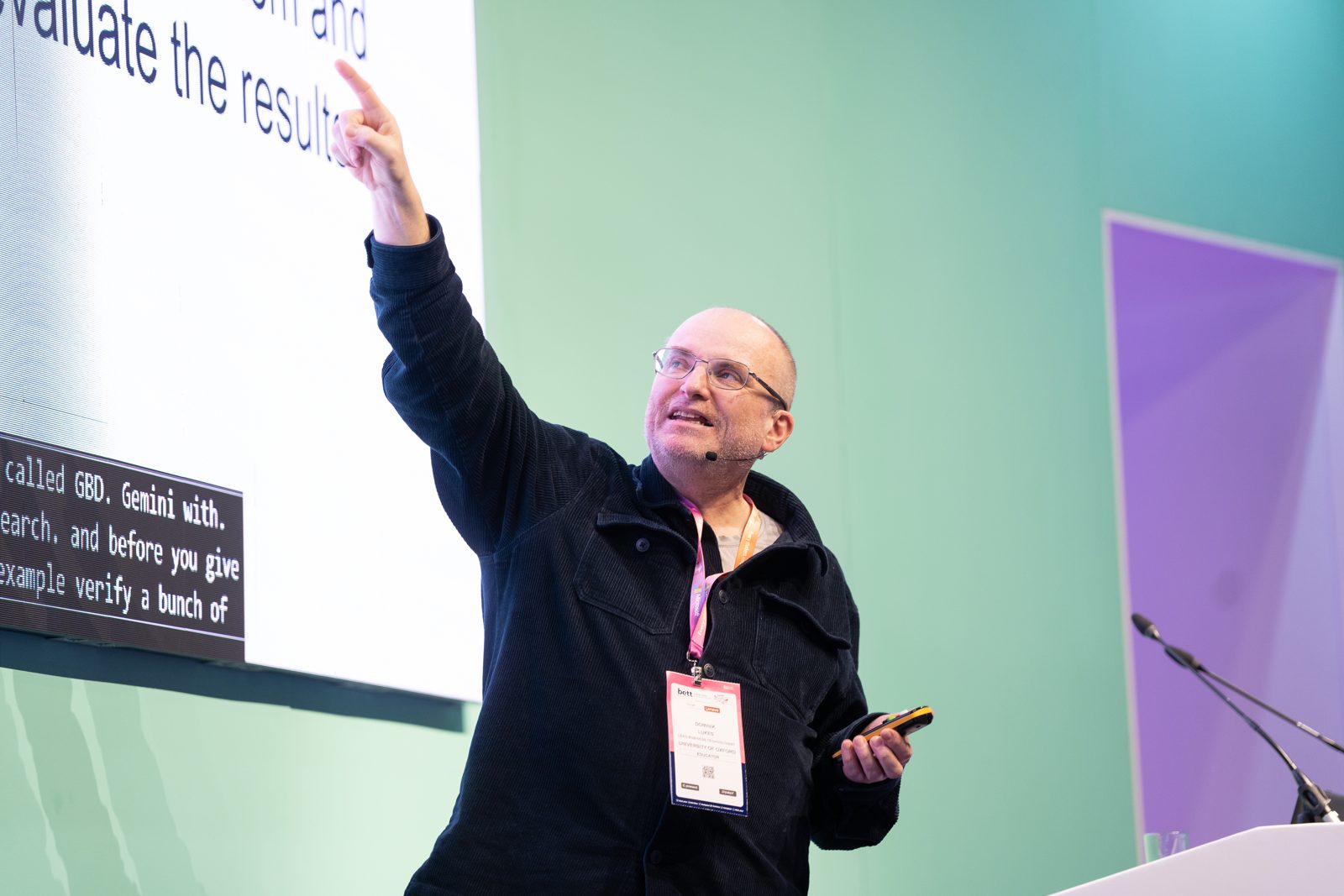 Dominik Lukeš on stage at Bett 2025, pointing up towards a large slide whose visible text includes “evaluate the results”